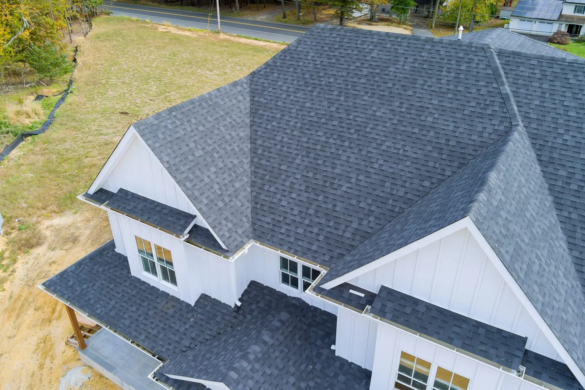 Aerial view of a large house with a dark gray shingle roof and white exterior, recently benefiting from residential roof replacement. The home boasts multiple gables and small windows, set on a grassy plot with a road and more houses in the background. Dominion Roofing | BRAMPTON | BURLINGTON | ETOBICOKE | MARKHAM | MISSISSAUGA | NORTH YORK | OAKVILLE | RICHMOND HILL | Scarborough | TORONTO | VAUGHAN | dominionroofing.com | Phone Number: 416-789-0601