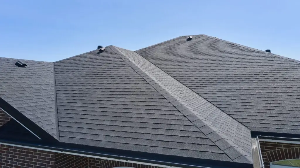 The image shows a close-up view of a multi-pitched roof with gray asphalt shingles on a clear day. Black vents are evenly spaced across the roof, and sections of brick walls are visible below the eaves. Dominion Roofing | BRAMPTON | BURLINGTON | ETOBICOKE | MARKHAM | MISSISSAUGA | NORTH YORK | OAKVILLE | RICHMOND HILL | Scarborough | TORONTO | VAUGHAN | dominionroofing.com | Phone Number: 416-789-0601