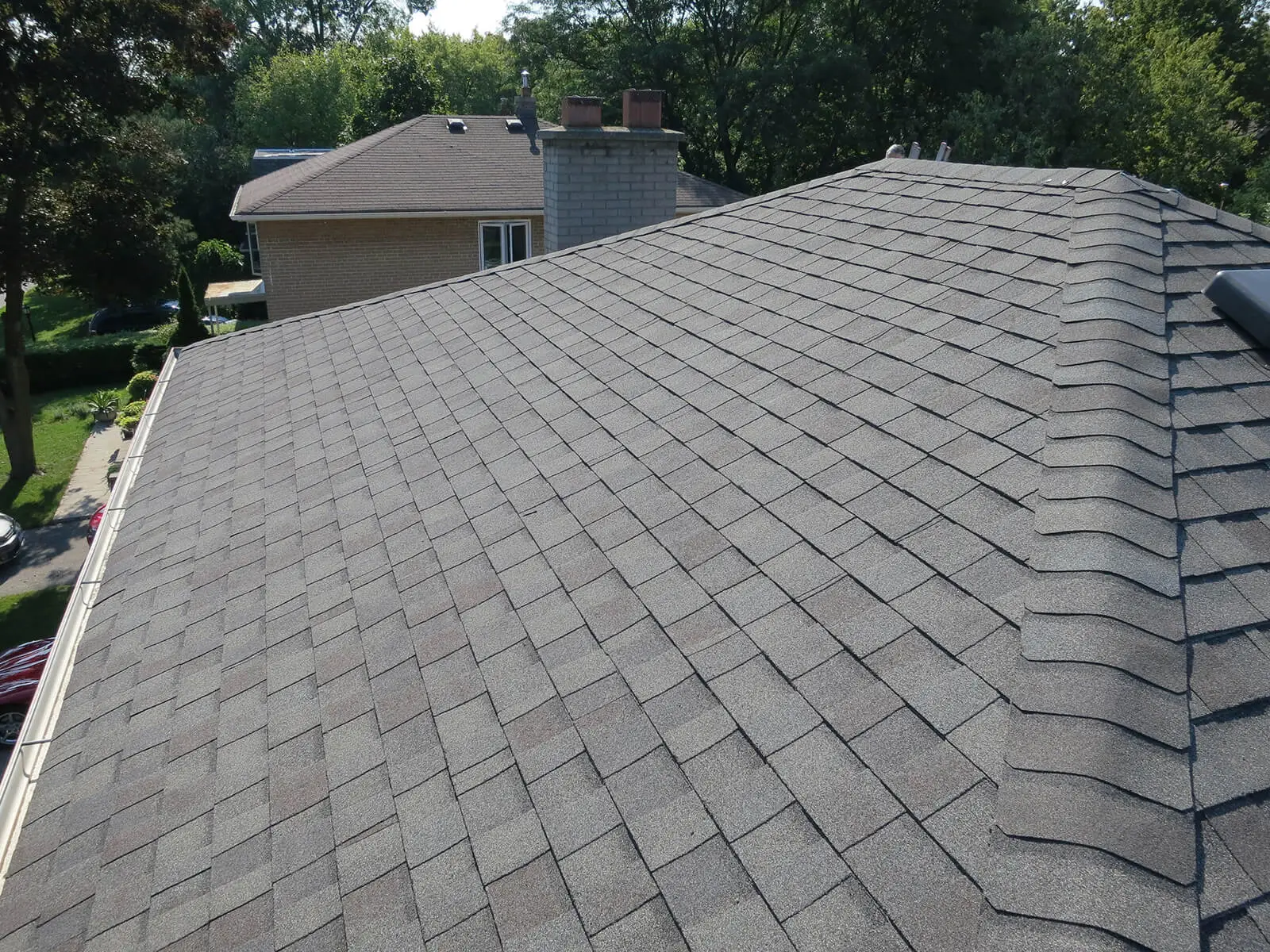 A view of a gray shingle roof extending across the image, surrounded by trees and a neighboring house in the background. The roof is clean and has a uniform pattern, with sunlight casting slight shadows on the shingles. Dominion Roofing | BRAMPTON | BURLINGTON | ETOBICOKE | MARKHAM | MISSISSAUGA | NORTH YORK | OAKVILLE | RICHMOND HILL | Scarborough | TORONTO | VAUGHAN | dominionroofing.com | Phone Number: 416-789-0601