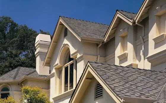 A close-up view of the roof and upper part of a beige house with dark grey residential shingle roofing. The house features gables, a large arched window, and a prominent chimney. Theres a clear blue sky and a tree partially visible in the background. Dominion Roofing | BRAMPTON | BURLINGTON | ETOBICOKE | MARKHAM | MISSISSAUGA | NORTH YORK | OAKVILLE | RICHMOND HILL | Scarborough | TORONTO | VAUGHAN | dominionroofing.com | Phone Number: 416-789-0601