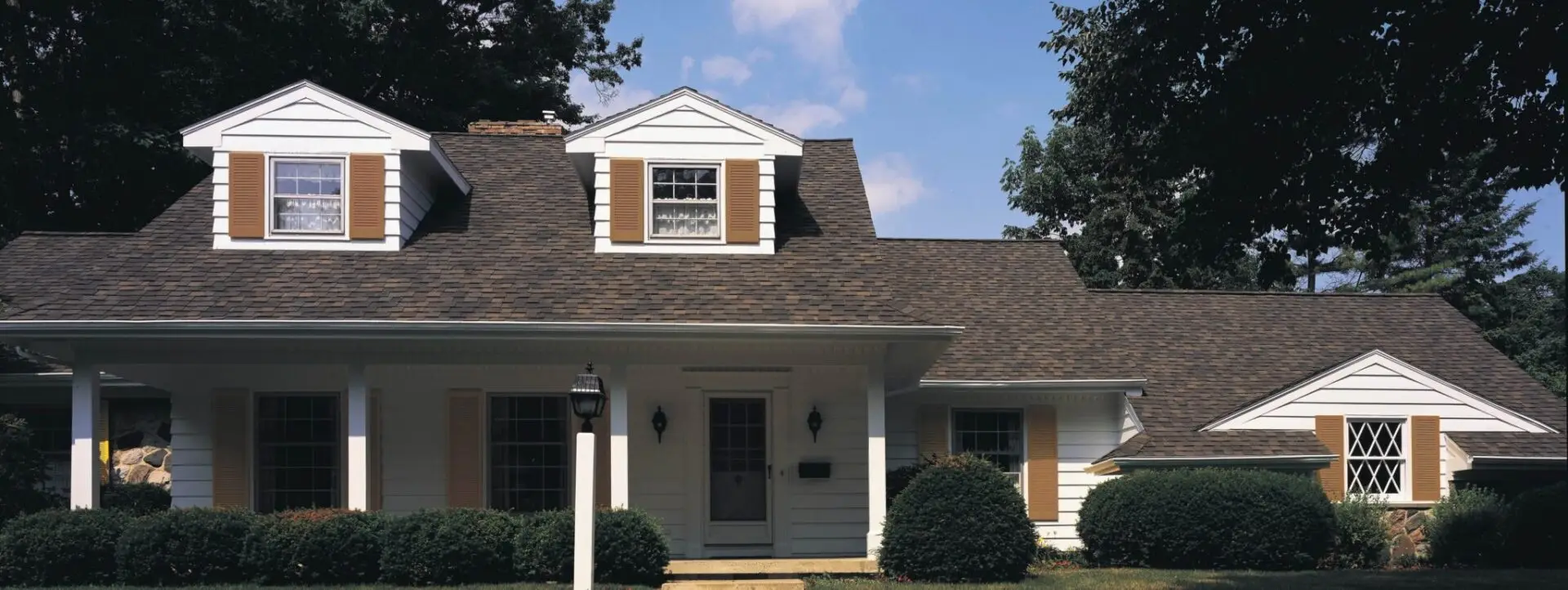 A white suburban house with a dark shingle roof and two dormer windows stands amidst green shrubs and trees. The front features a porch with columns and a central entrance with a glass door. A lamp post is on the front lawn, under a blue sky. Dominion Roofing | BRAMPTON | BURLINGTON | ETOBICOKE | MARKHAM | MISSISSAUGA | NORTH YORK | OAKVILLE | RICHMOND HILL | Scarborough | TORONTO | VAUGHAN | dominionroofing.com | Phone Number: 416-789-0601