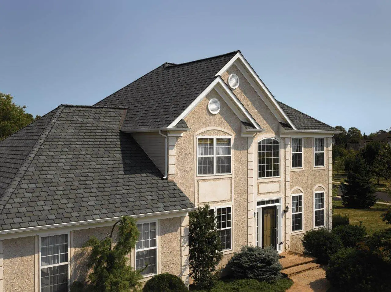 A two-story house with light-colored stucco walls and a dark gray shingle roof. It has large arched windows, a central entrance with pillars, and manicured shrubs and trees lining the pathway leading to the door. Dominion Roofing | BRAMPTON | BURLINGTON | ETOBICOKE | MARKHAM | MISSISSAUGA | NORTH YORK | OAKVILLE | RICHMOND HILL | Scarborough | TORONTO | VAUGHAN | dominionroofing.com | Phone Number: 416-789-0601