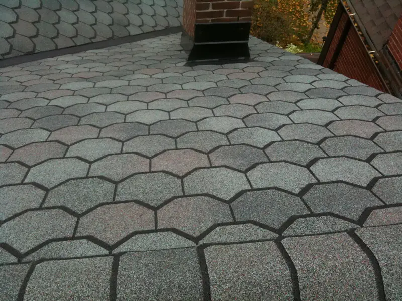 A roof covered with gray, hexagonal residential shingle roofing features asphalt shingles. The shingles have subtle color variations and create a textured pattern. A brick chimney rises from the roof, and some foliage is visible in the background. Dominion Roofing | BRAMPTON | BURLINGTON | ETOBICOKE | MARKHAM | MISSISSAUGA | NORTH YORK | OAKVILLE | RICHMOND HILL | Scarborough | TORONTO | VAUGHAN | dominionroofing.com | Phone Number: 416-789-0601
