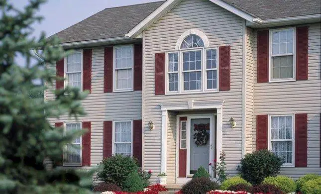 A two-story house with beige siding and red shutters showcases its residential shingle roofing. It features a large arched window above the front door, which is adorned with a wreath. The front yard is landscaped with bushes and flowering plants, while a tree is partially visible on the left. Dominion Roofing | BRAMPTON | BURLINGTON | ETOBICOKE | MARKHAM | MISSISSAUGA | NORTH YORK | OAKVILLE | RICHMOND HILL | Scarborough | TORONTO | VAUGHAN | dominionroofing.com | Phone Number: 416-789-0601