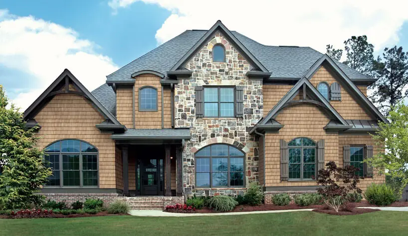 A two-story house featuring a mix of stone and wood-paneled exterior with sleek residential shingle roofing. The design includes multiple gables, arched windows, and a well-maintained lawn with shrubs. The sky is partly cloudy. Dominion Roofing | BRAMPTON | BURLINGTON | ETOBICOKE | MARKHAM | MISSISSAUGA | NORTH YORK | OAKVILLE | RICHMOND HILL | Scarborough | TORONTO | VAUGHAN | dominionroofing.com | Phone Number: 416-789-0601