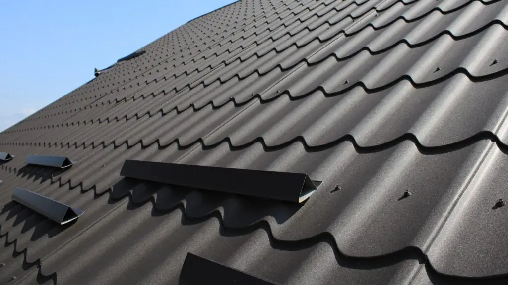 Close-up of a rooftop covered with dark metal tiles. The tiles have a wavy pattern, and there are several snow guards attached. The sky is clear and blue in the background. Dominion Roofing | BRAMPTON | BURLINGTON | ETOBICOKE | MARKHAM | MISSISSAUGA | NORTH YORK | OAKVILLE | RICHMOND HILL | Scarborough | TORONTO | VAUGHAN | dominionroofing.com | Phone Number: 416-789-0601