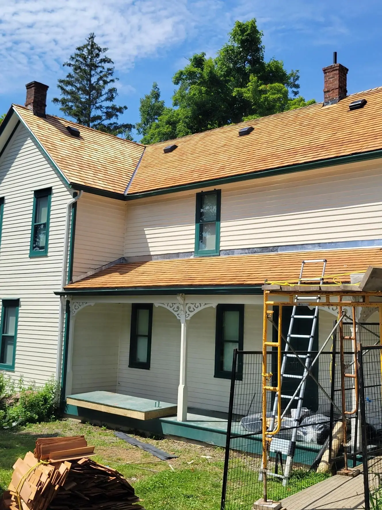 A two-story house with a yellowish-brown roof and light siding showcases its specialty residential roofing. Scaffolding is set up in front, with a ladder leaning against the roof. Bundles of roofing materials lie on the grass, framed by trees under a partly cloudy sky. Dominion Roofing | BRAMPTON | BURLINGTON | ETOBICOKE | MARKHAM | MISSISSAUGA | NORTH YORK | OAKVILLE | RICHMOND HILL | Scarborough | TORONTO | VAUGHAN | dominionroofing.com | Phone Number: 416-789-0601