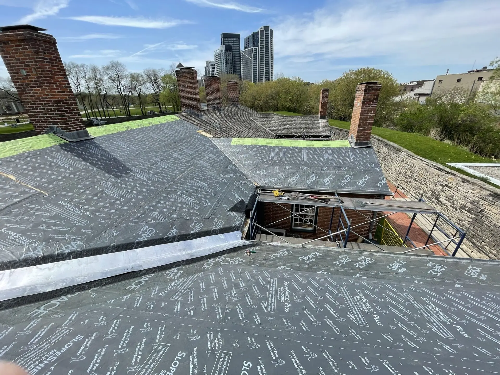 Witness a rooftop under construction, showcasing specialty commercial roofing solutions with sleek black sheathing material. Brick chimneys rise amidst the surrounding greenery, while high-rise buildings silhouette against a partly cloudy sky in the background. Dominion Roofing | BRAMPTON | BURLINGTON | ETOBICOKE | MARKHAM | MISSISSAUGA | NORTH YORK | OAKVILLE | RICHMOND HILL | Scarborough | TORONTO | VAUGHAN | dominionroofing.com | Phone Number: 416-789-0601