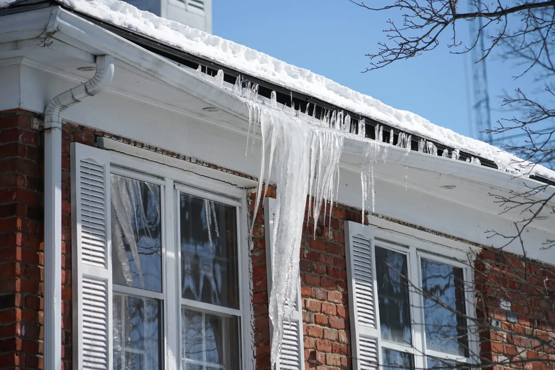 Large icicles hang from the snow-covered roof of a red brick house with white shutters, hinting at the need for a roof repair. The sky is clear and blue, with a few tree branches visible in the foreground. Dominion Roofing | BRAMPTON | BURLINGTON | ETOBICOKE | MARKHAM | MISSISSAUGA | NORTH YORK | OAKVILLE | RICHMOND HILL | Scarborough | TORONTO | VAUGHAN | dominionroofing.com | Phone Number: 416-789-0601
