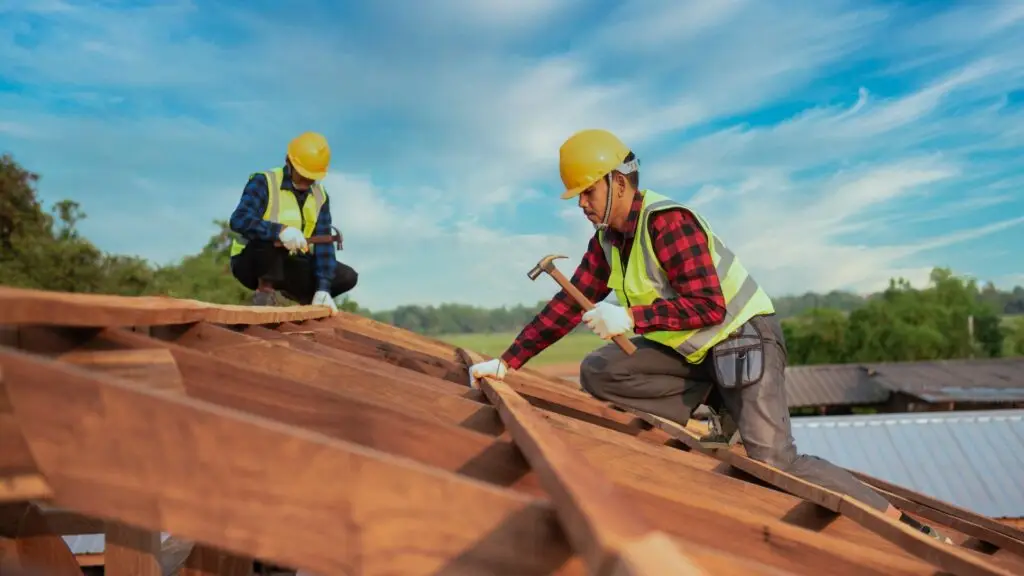 Two construction workers wearing yellow hard hats and reflective vests work on a wooden roof. One is hammering a nail, while the other holds a beam. The sky is clear and blue with scattered clouds in the background. Dominion Roofing | BRAMPTON | BURLINGTON | ETOBICOKE | MARKHAM | MISSISSAUGA | NORTH YORK | OAKVILLE | RICHMOND HILL | Scarborough | TORONTO | VAUGHAN | dominionroofing.com | Phone Number: 416-789-0601