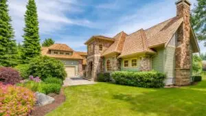 A charming suburban house with stone and wood exterior, surrounded by a well-manicured lawn, vibrant bushes, and tall trees. A clear blue sky is in the background, enhancing the scenes tranquility. Dominion Roofing | BRAMPTON | BURLINGTON | ETOBICOKE | MARKHAM | MISSISSAUGA | NORTH YORK | OAKVILLE | RICHMOND HILL | Scarborough | TORONTO | VAUGHAN | dominionroofing.com | Phone Number: 416-789-0601