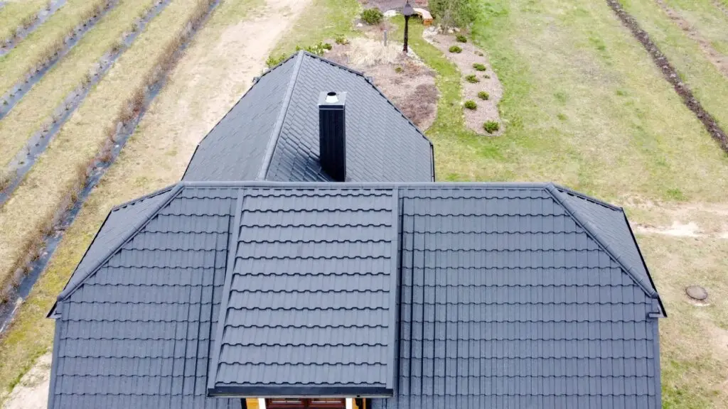 Aerial view of a house with a dark, textured metal roof. The house is surrounded by a garden with small plants and paths. The area around the house has rows of tilled soil, possibly for planting. Dominion Roofing | BRAMPTON | BURLINGTON | ETOBICOKE | MARKHAM | MISSISSAUGA | NORTH YORK | OAKVILLE | RICHMOND HILL | Scarborough | TORONTO | VAUGHAN | dominionroofing.com | Phone Number: 416-789-0601