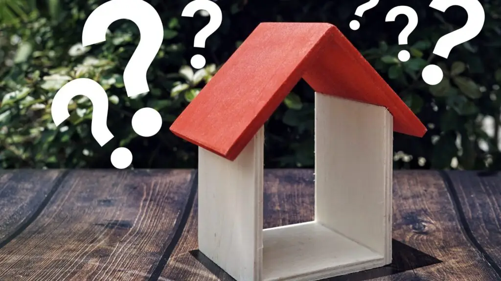 A small model wooden house with a red roof stands on a wooden surface. Surrounding it are multiple white question marks. Blurred greenery is in the background. Dominion Roofing | BRAMPTON | BURLINGTON | ETOBICOKE | MARKHAM | MISSISSAUGA | NORTH YORK | OAKVILLE | RICHMOND HILL | Scarborough | TORONTO | VAUGHAN | dominionroofing.com | Phone Number: 416-789-0601