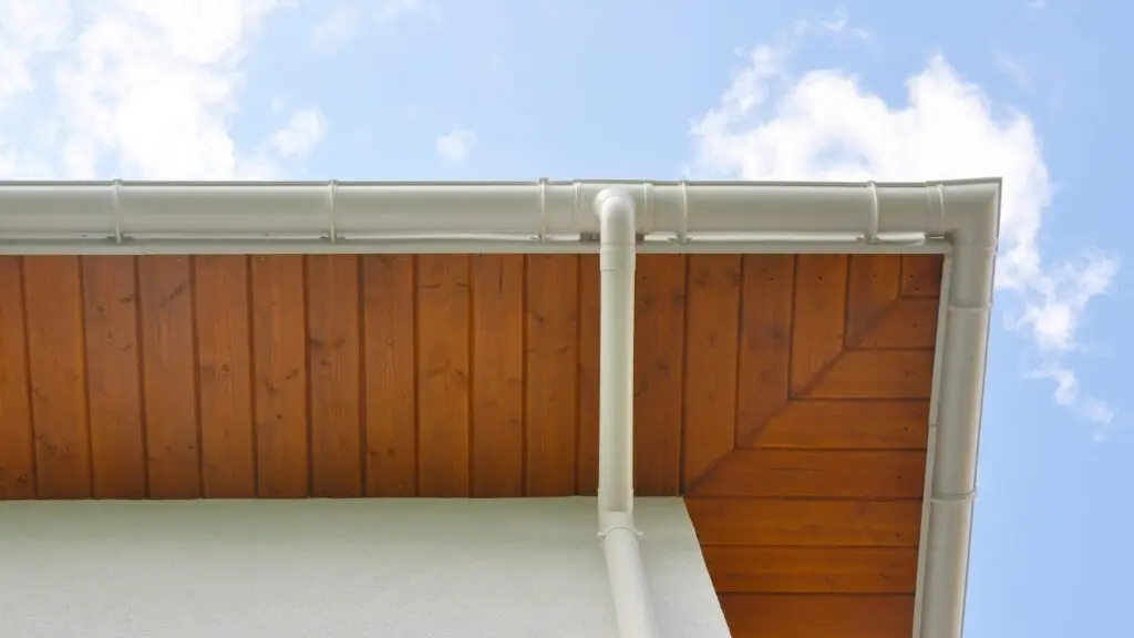 Close-up view of the corner of a building roof. The roof has light-colored gutters against a wooden soffit with a diagonal pattern. The background features a clear blue sky with a few clouds. Dominion Roofing | BRAMPTON | BURLINGTON | ETOBICOKE | MARKHAM | MISSISSAUGA | NORTH YORK | OAKVILLE | RICHMOND HILL | Scarborough | TORONTO | VAUGHAN | dominionroofing.com | Phone Number: 416-789-0601