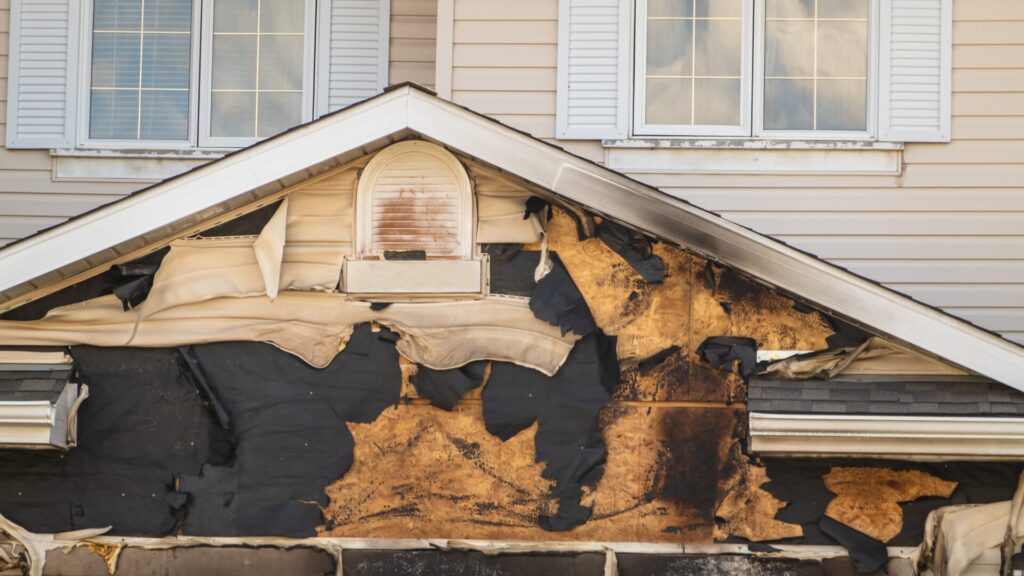 Damaged house facade with peeling siding and exposed, burnt materials. The windows above are intact, contrasting with the deteriorated section below. Dominion Roofing | BRAMPTON | BURLINGTON | ETOBICOKE | MARKHAM | MISSISSAUGA | NORTH YORK | OAKVILLE | RICHMOND HILL | Scarborough | TORONTO | VAUGHAN | dominionroofing.com | Phone Number: 416-789-0601