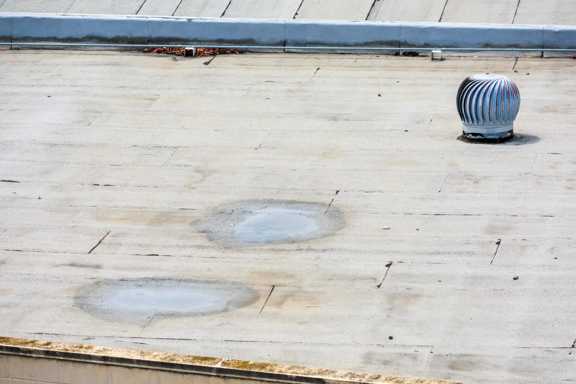 A flat rooftop with a metal turbine vent on the right side showcases the durability of concrete, featuring circular patches and light debris near the top. The roof edge is bordered by a light-colored parapet wall, exemplifying optimal Commercial Roof Replacement Solutions. Dominion Roofing | BRAMPTON | BURLINGTON | ETOBICOKE | MARKHAM | MISSISSAUGA | NORTH YORK | OAKVILLE | RICHMOND HILL | Scarborough | TORONTO | VAUGHAN | dominionroofing.com | Phone Number: 416-789-0601
