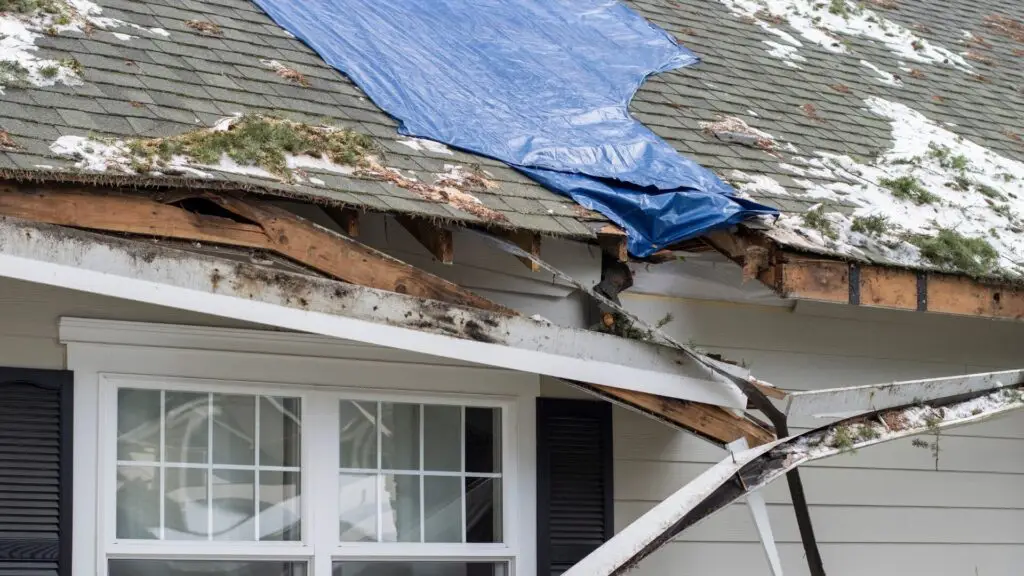 Damaged house roof with missing shingles and exposed wood, partially covered by a blue tarp. Snow and debris are scattered on the roof. A bent white support beam is visible above a window with black shutters. Dominion Roofing | BRAMPTON | BURLINGTON | ETOBICOKE | MARKHAM | MISSISSAUGA | NORTH YORK | OAKVILLE | RICHMOND HILL | Scarborough | TORONTO | VAUGHAN | dominionroofing.com | Phone Number: 416-789-0601