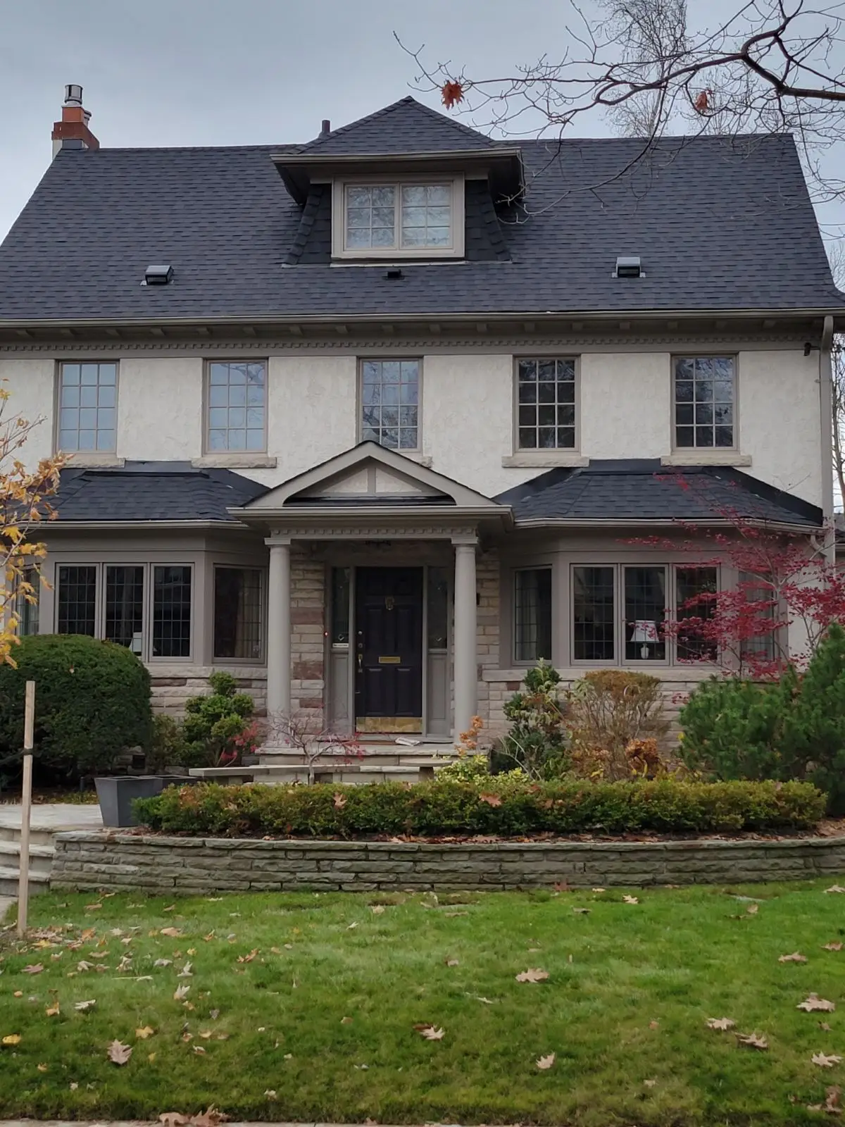 Two-story house with grayish-white exterior, dark residential shingle roofing, and a central black door. Symmetrical windows flank the entrance. A manicured lawn and shrubs enhance the front yard, with fallen leaves scattered on the grass. Overcast sky in the background. Dominion Roofing | BRAMPTON | BURLINGTON | ETOBICOKE | MARKHAM | MISSISSAUGA | NORTH YORK | OAKVILLE | RICHMOND HILL | Scarborough | TORONTO | VAUGHAN | dominionroofing.com | Phone Number: 416-789-0601