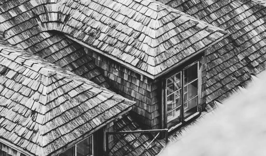 A black-and-white image showing a steeply pitched rooftop with wooden shingles. Two dormer windows protrude from the roof, framed in white, adding texture and depth to the rustic architectural scene. Dominion Roofing | BRAMPTON | BURLINGTON | ETOBICOKE | MARKHAM | MISSISSAUGA | NORTH YORK | OAKVILLE | RICHMOND HILL | Scarborough | TORONTO | VAUGHAN | dominionroofing.com | Phone Number: 416-789-0601