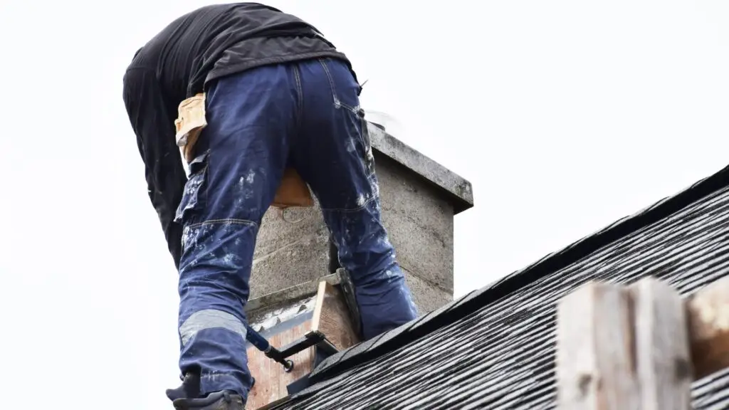 A person wearing a black jacket and blue paint-splattered pants is working on a rooftop, facing away from the camera. They are adjusting a wooden board near a chimney under a cloudy sky. The roof is covered with dark shingles. Dominion Roofing | BRAMPTON | BURLINGTON | ETOBICOKE | MARKHAM | MISSISSAUGA | NORTH YORK | OAKVILLE | RICHMOND HILL | Scarborough | TORONTO | VAUGHAN | dominionroofing.com | Phone Number: 416-789-0601