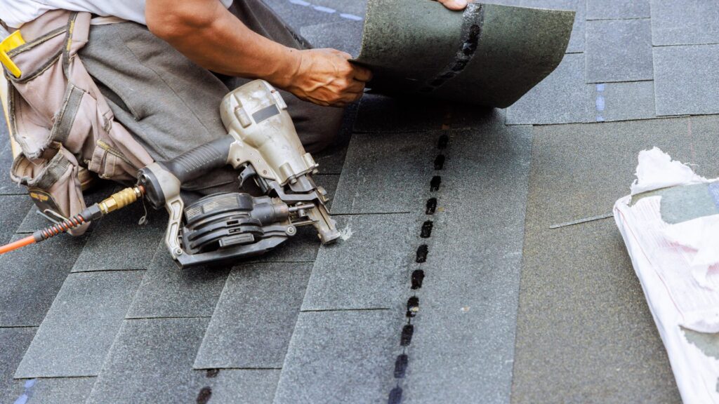 Worker laying asphalt shingles on a roof, holding a nail gun with several shingles already in place. The worker is wearing a tool belt and using black roofing nails in a precise row. Dominion Roofing | BRAMPTON | BURLINGTON | ETOBICOKE | MARKHAM | MISSISSAUGA | NORTH YORK | OAKVILLE | RICHMOND HILL | Scarborough | TORONTO | VAUGHAN | dominionroofing.com | Phone Number: 416-789-0601