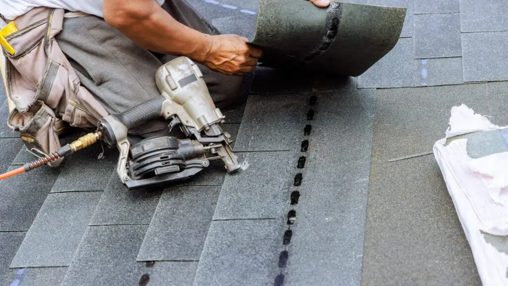 Worker laying asphalt shingles on a roof, holding a nail gun with several shingles already in place. The worker is wearing a tool belt and using black roofing nails in a precise row. Dominion Roofing | BRAMPTON | BURLINGTON | ETOBICOKE | MARKHAM | MISSISSAUGA | NORTH YORK | OAKVILLE | RICHMOND HILL | Scarborough | TORONTO | VAUGHAN | dominionroofing.com | Phone Number: 416-789-0601
