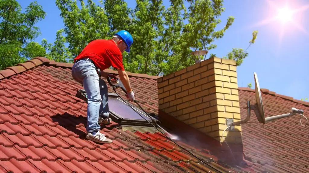 A worker in a red shirt and blue helmet is power washing a red tile roof near a yellow brick chimney. A satellite dish is mounted nearby. The sun shines brightly in a clear blue sky with green trees in the background. Dominion Roofing | BRAMPTON | BURLINGTON | ETOBICOKE | MARKHAM | MISSISSAUGA | NORTH YORK | OAKVILLE | RICHMOND HILL | Scarborough | TORONTO | VAUGHAN | dominionroofing.com | Phone Number: 416-789-0601