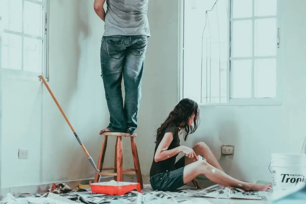 A man stands on a stool painting a wall with a roller, while a woman sits on the floor resting. The room is filled with natural light from large windows, and newspaper covers the floor to catch paint drips. Dominion Roofing | BRAMPTON | BURLINGTON | ETOBICOKE | MARKHAM | MISSISSAUGA | NORTH YORK | OAKVILLE | RICHMOND HILL | Scarborough | TORONTO | VAUGHAN | dominionroofing.com | Phone Number: 416-789-0601