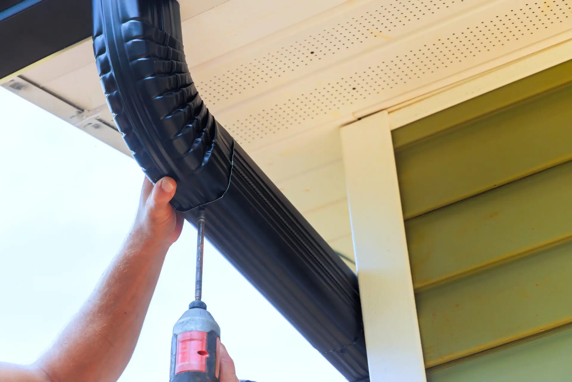 A person expertly uses a power drill to install a black downspout at the corner of a building with green siding, showcasing the precision of residential eavestrough and downpipe services. The view highlights the hands, drill, and downspout beneath a beige soffit. Dominion Roofing | BRAMPTON | BURLINGTON | ETOBICOKE | MARKHAM | MISSISSAUGA | NORTH YORK | OAKVILLE | RICHMOND HILL | Scarborough | TORONTO | VAUGHAN | dominionroofing.com | Phone Number: 416-789-0601