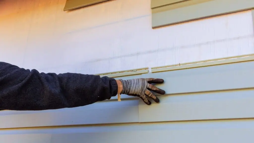 A persons gloved hand adjusts light blue vinyl siding on a house. The wall appears partially completed with a white area visible above. The person is wearing a dark long-sleeve top. Dominion Roofing | BRAMPTON | BURLINGTON | ETOBICOKE | MARKHAM | MISSISSAUGA | NORTH YORK | OAKVILLE | RICHMOND HILL | Scarborough | TORONTO | VAUGHAN | dominionroofing.com | Phone Number: 416-789-0601