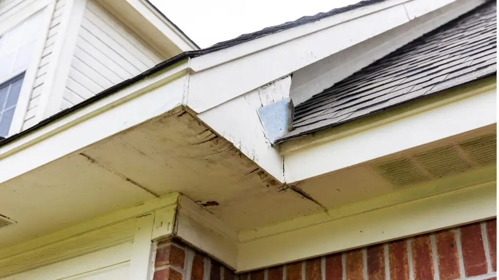 Close-up of a houses roof with visible water damage and rot on the wooden soffit and fascia boards. The bricks and white window frame of the house are partially visible below the gutter line. The sky is overcast. Dominion Roofing | BRAMPTON | BURLINGTON | ETOBICOKE | MARKHAM | MISSISSAUGA | NORTH YORK | OAKVILLE | RICHMOND HILL | Scarborough | TORONTO | VAUGHAN | dominionroofing.com | Phone Number: 416-789-0601