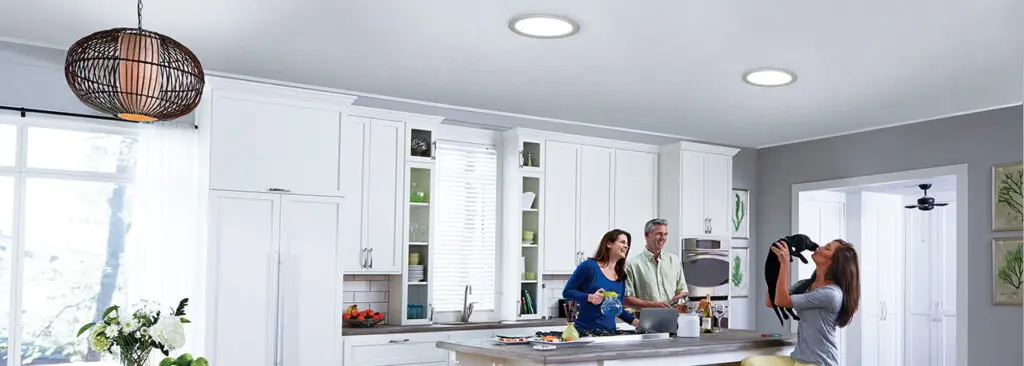 A modern kitchen with white cabinets and an island. Four people are gathered around, preparing food and drinks. A hanging light fixture is visible above, and the room is brightened by natural light through a window. Dominion Roofing | BRAMPTON | BURLINGTON | ETOBICOKE | MARKHAM | MISSISSAUGA | NORTH YORK | OAKVILLE | RICHMOND HILL | Scarborough | TORONTO | VAUGHAN | dominionroofing.com | Phone Number: 416-789-0601