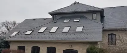 A large house with a sloped gray shingle roof featuring multiple skylights. The lower section of the building is constructed with light-colored stone. There are several bare trees and bushes surrounding the house under a cloudy sky. Dominion Roofing | BRAMPTON | BURLINGTON | ETOBICOKE | MARKHAM | MISSISSAUGA | NORTH YORK | OAKVILLE | RICHMOND HILL | Scarborough | TORONTO | VAUGHAN | dominionroofing.com | Phone Number: 416-789-0601