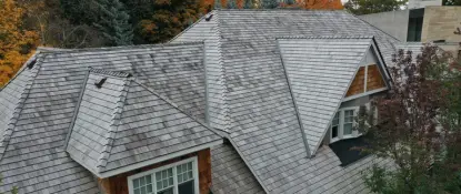 A rooftop with weathered wooden shingles and a contemporary design. It features triangular gables and multiple peaks, surrounded by autumn foliage with trees showing green and orange leaves. Dominion Roofing | BRAMPTON | BURLINGTON | ETOBICOKE | MARKHAM | MISSISSAUGA | NORTH YORK | OAKVILLE | RICHMOND HILL | Scarborough | TORONTO | VAUGHAN | dominionroofing.com | Phone Number: 416-789-0601