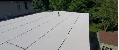 Flat rooftop with a white surface and dark lines, surrounded by greenery and parts of neighboring buildings visible. The sky is clear and sunlight casts shadows on the roof. Dominion Roofing | BRAMPTON | BURLINGTON | ETOBICOKE | MARKHAM | MISSISSAUGA | NORTH YORK | OAKVILLE | RICHMOND HILL | Scarborough | TORONTO | VAUGHAN | dominionroofing.com | Phone Number: 416-789-0601