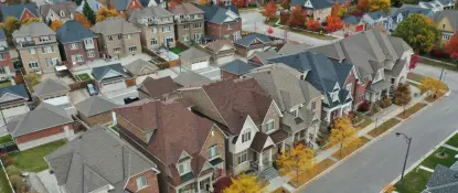 Aerial view of a suburban neighborhood with rows of similar two-story houses. Trees with autumn foliage line the streets. The houses have varied roof styles and colors, creating a patchwork appearance. The streets are empty, indicating a quiet area. Dominion Roofing | BRAMPTON | BURLINGTON | ETOBICOKE | MARKHAM | MISSISSAUGA | NORTH YORK | OAKVILLE | RICHMOND HILL | Scarborough | TORONTO | VAUGHAN | dominionroofing.com | Phone Number: 416-789-0601