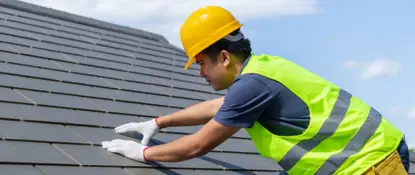 A construction worker wearing a yellow hard hat, a reflective vest, and white gloves installs shingles on a roof under a clear blue sky. Dominion Roofing | BRAMPTON | BURLINGTON | ETOBICOKE | MARKHAM | MISSISSAUGA | NORTH YORK | OAKVILLE | RICHMOND HILL | Scarborough | TORONTO | VAUGHAN | dominionroofing.com | Phone Number: 416-789-0601