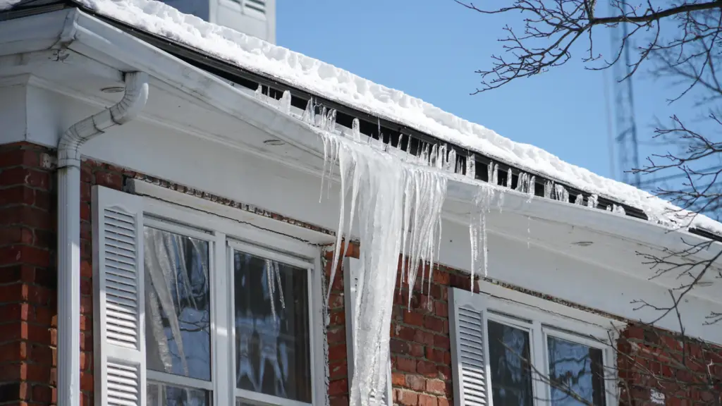 A brick house with large icicles hanging from a snow-covered roof. The icicles extend down past the windows. Bare tree branches are visible against a clear blue sky. Dominion Roofing | BRAMPTON | BURLINGTON | ETOBICOKE | MARKHAM | MISSISSAUGA | NORTH YORK | OAKVILLE | RICHMOND HILL | Scarborough | TORONTO | VAUGHAN | dominionroofing.com | Phone Number: 416-789-0601