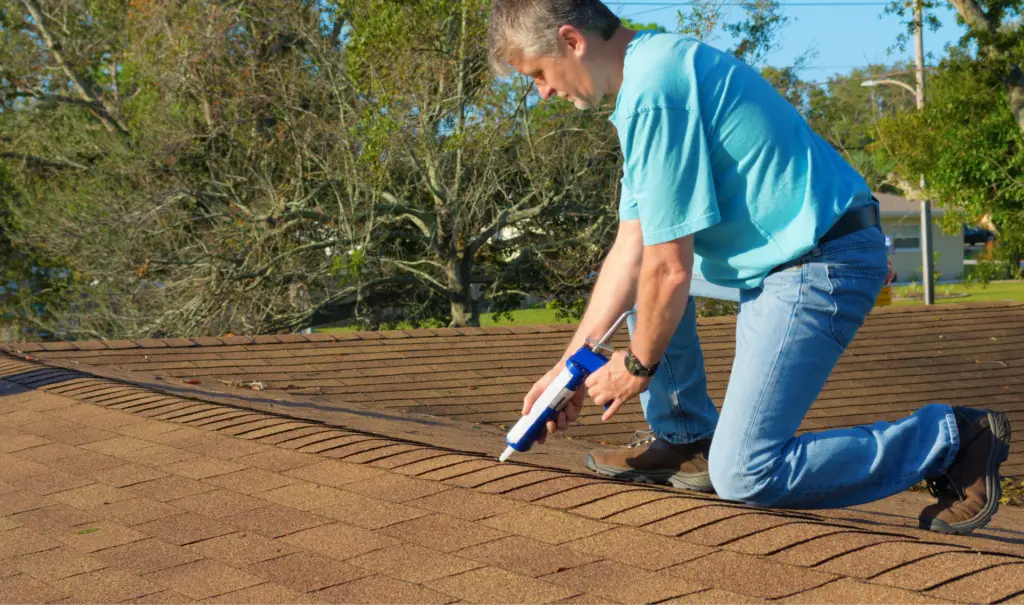 A person in a blue shirt and jeans kneels on a shingled roof, using a caulking gun to apply sealant along the ridge. Trees and a clear sky are in the background. Dominion Roofing | BRAMPTON | BURLINGTON | ETOBICOKE | MARKHAM | MISSISSAUGA | NORTH YORK | OAKVILLE | RICHMOND HILL | Scarborough | TORONTO | VAUGHAN | dominionroofing.com | Phone Number: 416-789-0601