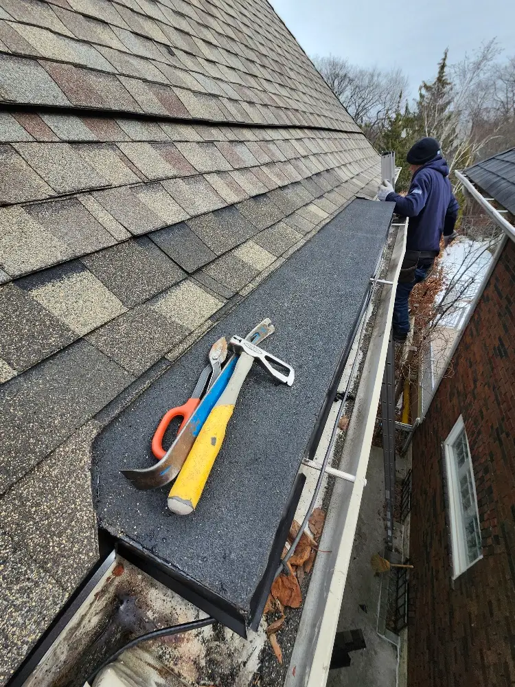 A person in a hoodie stands on a ladder next to a house roof, with three tools—a hammer, a pry bar, and snips—lying on black roofing material near the gutter, surrounded by shingles and fallen leaves. Dominion Roofing | BRAMPTON | BURLINGTON | ETOBICOKE | MARKHAM | MISSISSAUGA | NORTH YORK | OAKVILLE | RICHMOND HILL | Scarborough | TORONTO | VAUGHAN | dominionroofing.com | Phone Number: 416-789-0601