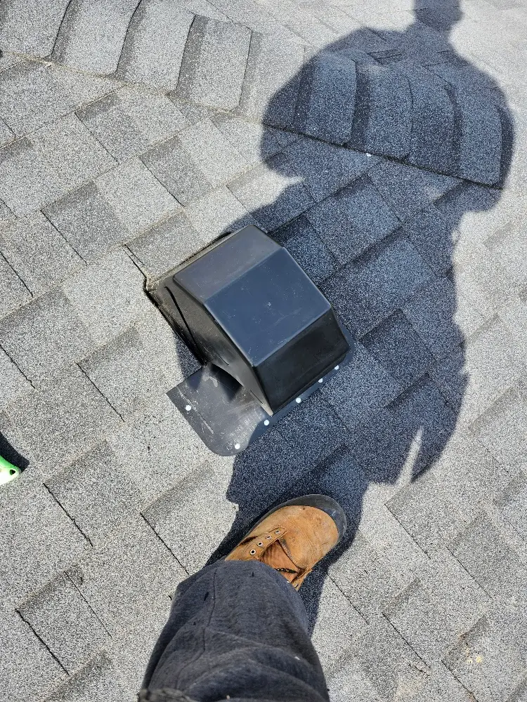 A person wearing a brown work boot stands on a gray shingled roof next to a black roof vent; the persons shadow is visible on the shingles. Dominion Roofing | BRAMPTON | BURLINGTON | ETOBICOKE | MARKHAM | MISSISSAUGA | NORTH YORK | OAKVILLE | RICHMOND HILL | Scarborough | TORONTO | VAUGHAN | dominionroofing.com | Phone Number: 416-789-0601