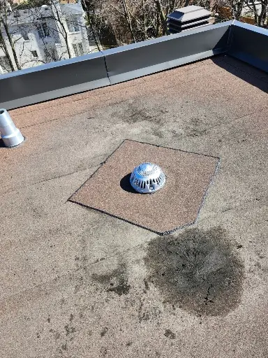 A flat rooftop with a metal drain cover in the center, surrounded by a patch of differently colored roofing material and a dark stain nearby. A vent pipe and a parapet wall are visible in the background. Dominion Roofing | BRAMPTON | BURLINGTON | ETOBICOKE | MARKHAM | MISSISSAUGA | NORTH YORK | OAKVILLE | RICHMOND HILL | Scarborough | TORONTO | VAUGHAN | dominionroofing.com | Phone Number: 416-789-0601