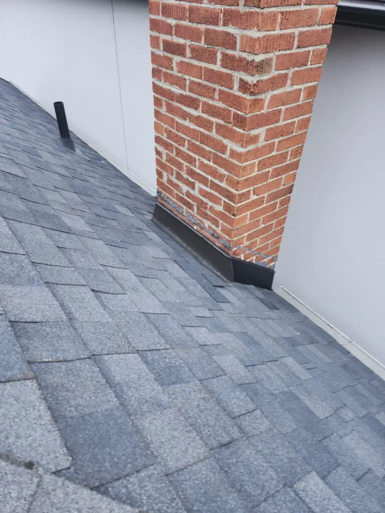 Gray asphalt shingles on a sloped roof with a red brick chimney and metal flashing at the base, next to a white wall. Dominion Roofing | BRAMPTON | BURLINGTON | ETOBICOKE | MARKHAM | MISSISSAUGA | NORTH YORK | OAKVILLE | RICHMOND HILL | Scarborough | TORONTO | VAUGHAN | dominionroofing.com | Phone Number: 416-789-0601