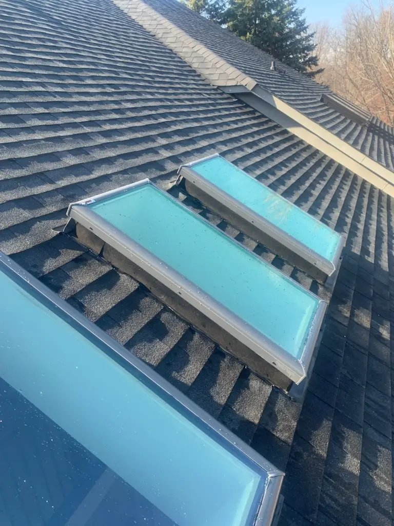 Three rectangular skylights set into a dark gray shingled roof, with trees and clear sky visible in the background. The skylights have a blue tint and are aligned in a row. Dominion Roofing | BRAMPTON | BURLINGTON | ETOBICOKE | MARKHAM | MISSISSAUGA | NORTH YORK | OAKVILLE | RICHMOND HILL | Scarborough | TORONTO | VAUGHAN | dominionroofing.com | Phone Number: 416-789-0601