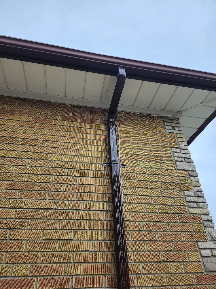 A close-up of a houses exterior shows a brown downspout attached to a yellow brick wall with some stonework on the corner, under a roof with white soffit and dark brown guttering against a cloudy sky. Dominion Roofing | BRAMPTON | BURLINGTON | ETOBICOKE | MARKHAM | MISSISSAUGA | NORTH YORK | OAKVILLE | RICHMOND HILL | Scarborough | TORONTO | VAUGHAN | dominionroofing.com | Phone Number: 416-789-0601