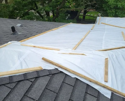 A rooftop with gray shingles, undergoing roof repair, is covered with large plastic sheets held down by wooden planks. The background showcases lush green trees and part of a neighboring house. Dominion Roofing | BRAMPTON | BURLINGTON | ETOBICOKE | MARKHAM | MISSISSAUGA | NORTH YORK | OAKVILLE | RICHMOND HILL | Scarborough | TORONTO | VAUGHAN | dominionroofing.com | Phone Number: 416-789-0601