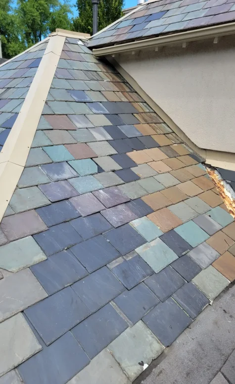 A multicolored slate roof, showcasing specialty residential roofing with shades of blue, green, and brown tiles. It features a steep slope and a section of rusted metal flashing. In the background, part of a chimney and neighboring roofs add to the unique character. Dominion Roofing | BRAMPTON | BURLINGTON | ETOBICOKE | MARKHAM | MISSISSAUGA | NORTH YORK | OAKVILLE | RICHMOND HILL | Scarborough | TORONTO | VAUGHAN | dominionroofing.com | Phone Number: 416-789-0601