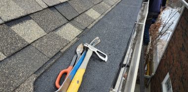 A person in a hoodie stands on a ladder next to a house roof, with three tools—a hammer, a pry bar, and snips—lying on black roofing material near the gutter, surrounded by shingles and fallen leaves. Dominion Roofing | BRAMPTON | BURLINGTON | ETOBICOKE | MARKHAM | MISSISSAUGA | NORTH YORK | OAKVILLE | RICHMOND HILL | Scarborough | TORONTO | VAUGHAN | dominionroofing.com | Phone Number: 416-789-0601