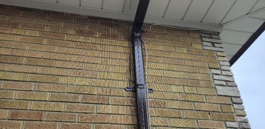 A close-up of a houses exterior shows a brown downspout attached to a yellow brick wall with some stonework on the corner, under a roof with white soffit and dark brown guttering against a cloudy sky. Dominion Roofing | BRAMPTON | BURLINGTON | ETOBICOKE | MARKHAM | MISSISSAUGA | NORTH YORK | OAKVILLE | RICHMOND HILL | Scarborough | TORONTO | VAUGHAN | dominionroofing.com | Phone Number: 416-789-0601