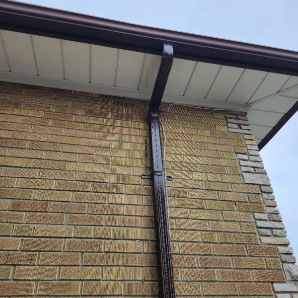 A close-up of a houses exterior shows a brown downspout attached to a yellow brick wall with some stonework on the corner, under a roof with white soffit and dark brown guttering against a cloudy sky. Dominion Roofing | BRAMPTON | BURLINGTON | ETOBICOKE | MARKHAM | MISSISSAUGA | NORTH YORK | OAKVILLE | RICHMOND HILL | Scarborough | TORONTO | VAUGHAN | dominionroofing.com | Phone Number: 416-789-0601